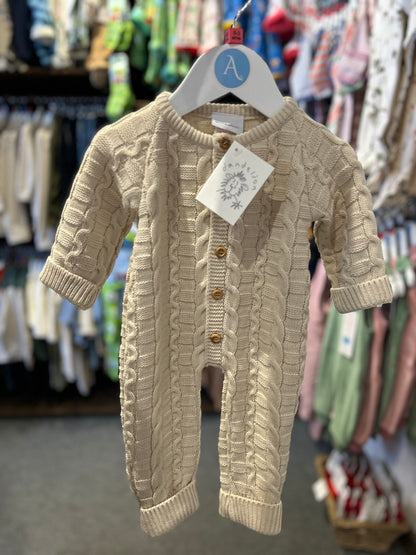 Dandelion basket weave knitted baby onesie in warm oatmeal taupe hanging on an Annabel's of Cranbrook branded white hanger with blue A roundel; photographed in the Annabel's of Cranbrook shop with colourful children's clothing rails softly blurred in the background; Dandelion swing tag hanging from the top button; full front view of the footless all-in-one showing the three-dimensional basket weave and cable knit texture in detail; five round gold-toned buttons down the centre front; ribbed neckband, sleeve