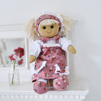 A blonde haired ragdoll wearing a blush pink floral corduroy dress with white lace trimmed pockets, a white long sleeved shirt with a blush pink floral print cord collar, and matching little pink corduroy shoes. The ragdoll is seated against a white background with a vase of red flowers in the background. BLONDE BLUSH FLOWER RAGDOLL WITH HEADBAND | POWELL CRAFT