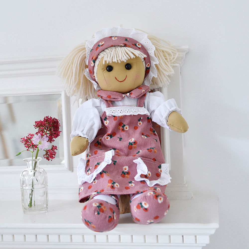 Toy doll in a floral outfit sitting on a white surface with a vase of flowers in the background. BLONDE BLUSH FLOWER RAGDOLL WITH HEADBAND | POWELL CRAFT