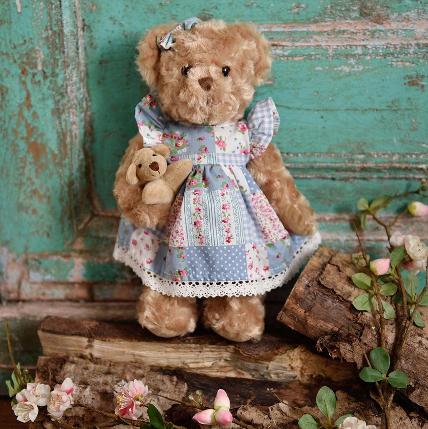 Teddy bear in a floral dress holding a smaller teddy bear against a rustic wooden background. BLUE FLORAL PATCH-WORK DRESS TEDDY & BABY | POWELL CRAFT