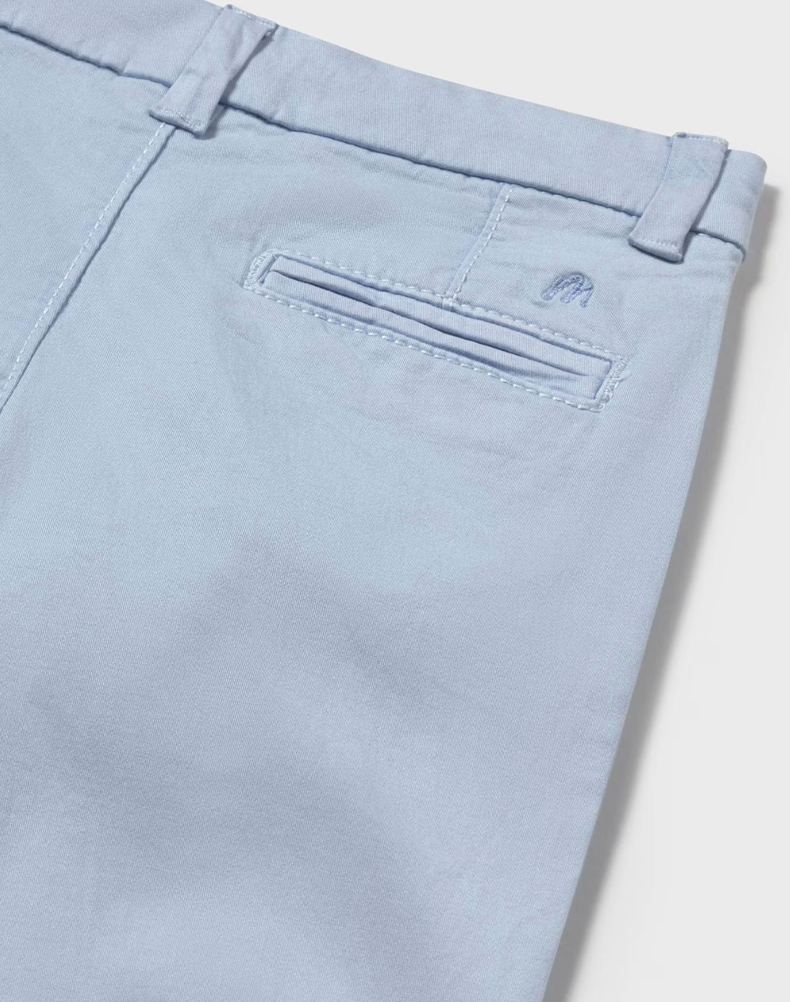  Mayoral Boys Basic Baby Blue Chino Trousers — close-up detail shot on white background; angled overhead view of the back right hip and waistband; powder/pale blue cotton-elastane fabric texture clearly visible with a fine chino weave; single welt/jetted back pocket shown in close detail with topstitched pocket opening; small tonal embroidered Mayoral stylised M logo in matching powder blue clearly visible just to the right of the pocket opening; belt loop at the right back waistband visible above; demonstr