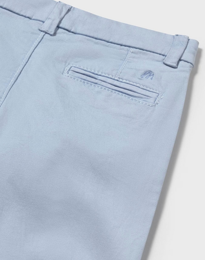  Mayoral Boys Basic Baby Blue Chino Trousers — close-up detail shot on white background; angled overhead view of the back right hip and waistband; powder/pale blue cotton-elastane fabric texture clearly visible with a fine chino weave; single welt/jetted back pocket shown in close detail with topstitched pocket opening; small tonal embroidered Mayoral stylised M logo in matching powder blue clearly visible just to the right of the pocket opening; belt loop at the right back waistband visible above; demonstr