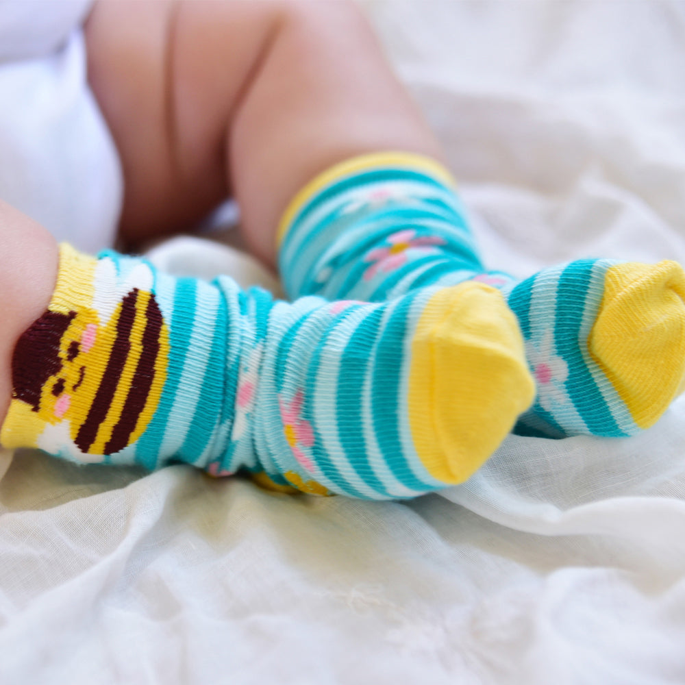 A pair of striped socks with contrasting yellow toes, heels, and top, decorated with pastel colored daisies and bees on the cuffs, on a baby's feet. CHILDREN'S BEE & BLUE STRIPE SOCKS - 3 SIZES | POWELL CRAFT