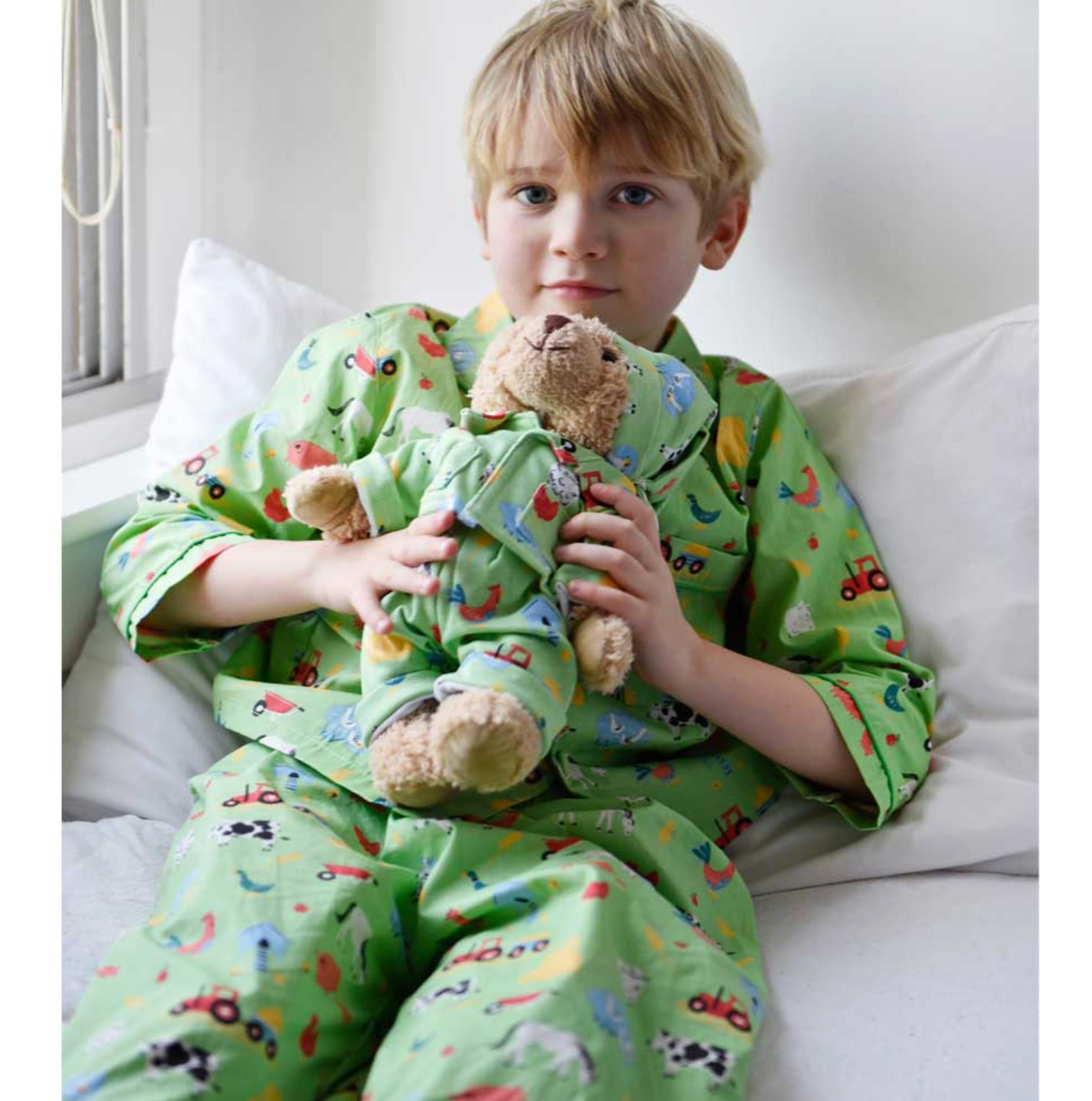 Child wearing green pajamas with vehicle prints, holding a MATCHING teddy bear on a white couch. CHILDREN'S CLASSIC BUTTON UP FARMYARD PYJAMAS | POWELL CRAFT