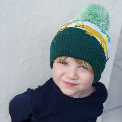 Child wearing a green knit hat with a colorful design and pom-pom. CHILDREN'S DINOSAUR KNITTED HAT WITH POM-POM | POWELL CRAFT