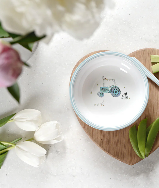 Small white bowl with blue rim and tractor design on a wooden coaster with tulips and flowers in the background. CHILDREN'S MELAMINE BOWL - ON THE FARM | SOPHIE ALLPORT