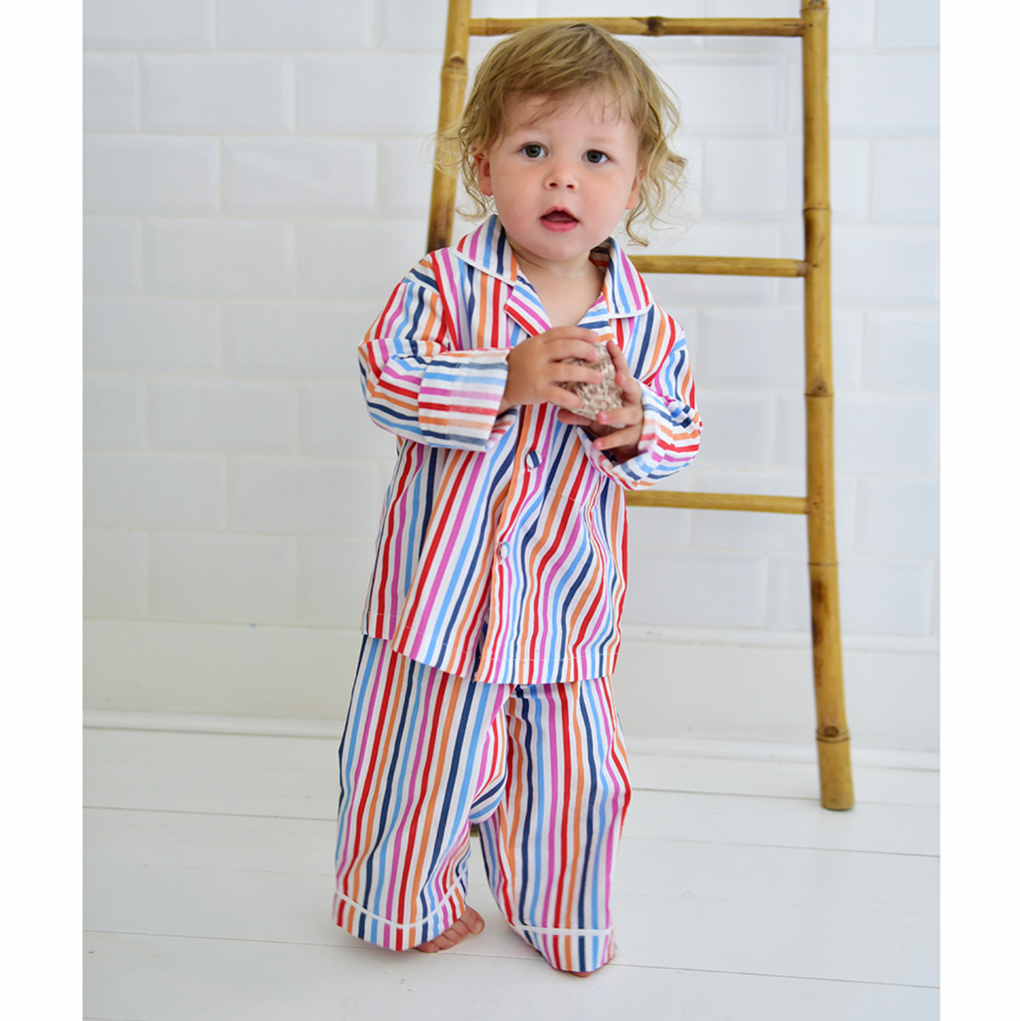 Child wearing a colorful striped outfit standing next to a wooden ladder on a white floor. CHILDREN'S MULTI-COLOURED STRIPE PYJAMAS | POWELL CRAFT