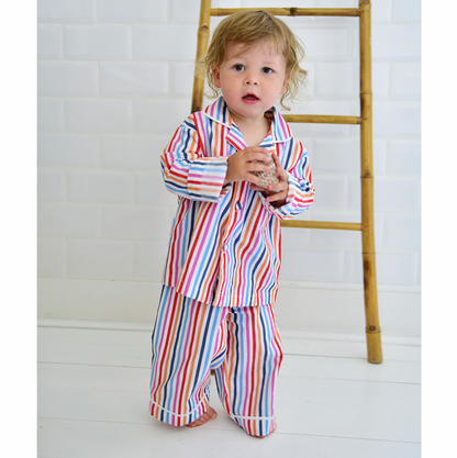 Child wearing a colorful striped outfit standing next to a wooden ladder on a white floor. CHILDREN'S MULTI-COLOURED STRIPE PYJAMAS | POWELL CRAFT