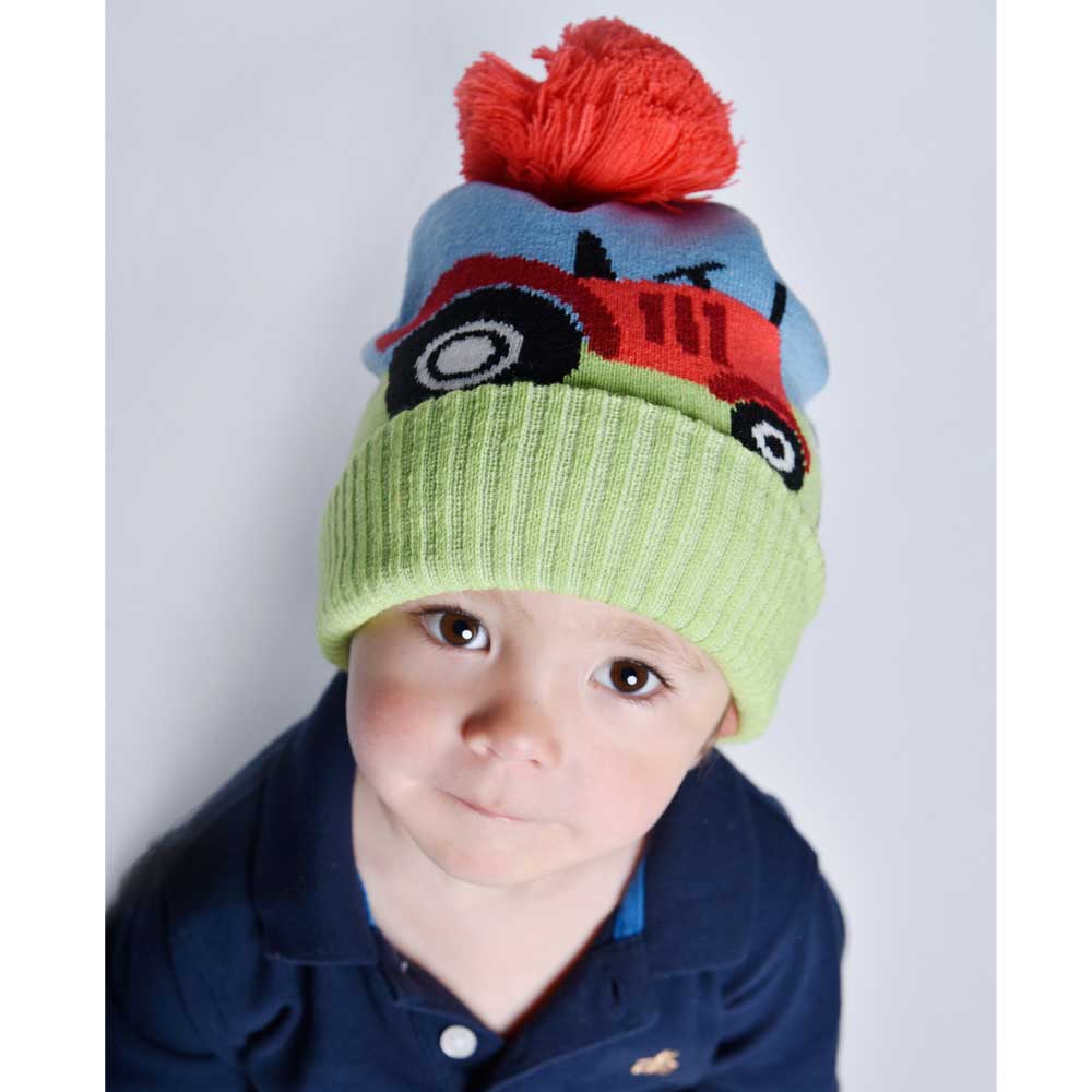 Child wearing a colorful knit hat with a red pom-pom and toy truck design. CHILDREN'S RED TRACTOR & SHEEP KNITTED HAT WITH POM-POM | POWELL CRAFT