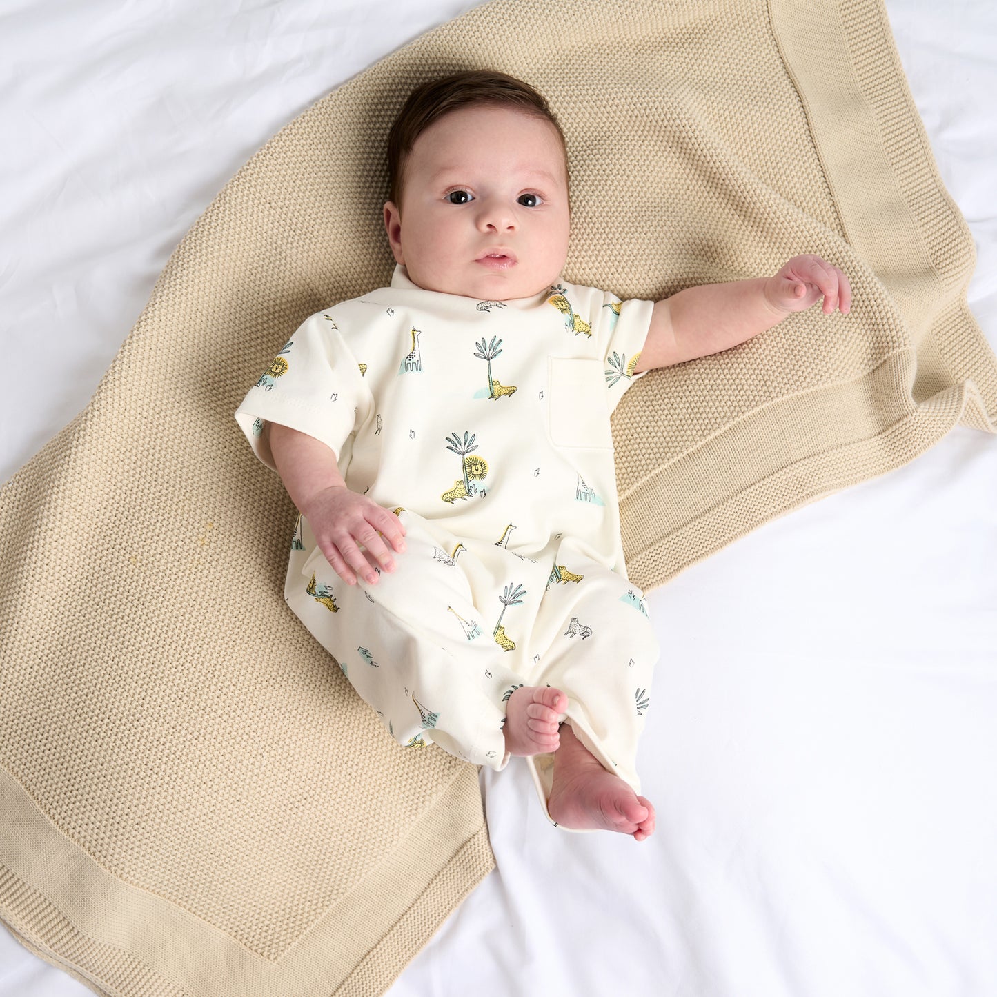 A baby lying on a beige blanket wearing a short-sleeved beige cotton romper with a jungle animal print. COTTON BABY ROMPER - JUNGLE FRIENDS | DANDELION