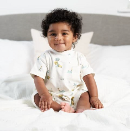 A baby sitting on a bed wearing a white short sleeve cotton romper with a jungle animal print and poppers at the back for easy dressing. COTTON BABY ROMPER - JUNGLE FRIENDS | DANDELION