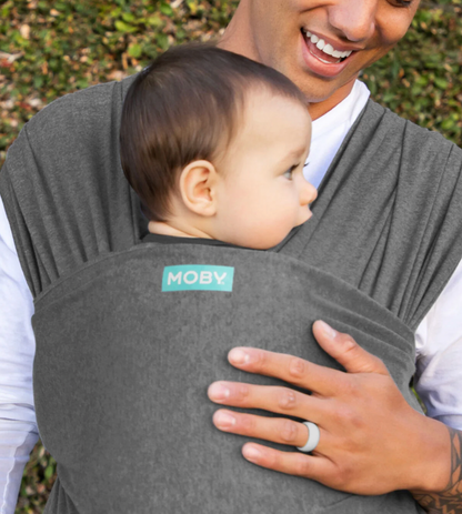 Person holding a baby in a gray Moby carrier outdoors. ELEMENTS SOFT & ADJUSTABLE FEATHER KNIT BABY WRAP CARRIER - 2 COLOURS | MOBY