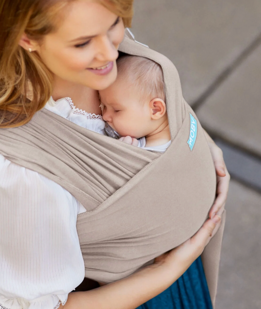 Woman holding a baby in a beige sling against a neutral background. ELEMENTS SOFT & ADJUSTABLE FEATHER KNIT BABY WRAP CARRIER - 2 COLOURS | MOBY