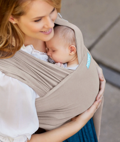 Woman holding a baby in a beige sling against a neutral background. ELEMENTS SOFT & ADJUSTABLE FEATHER KNIT BABY WRAP CARRIER - 2 COLOURS | MOBY