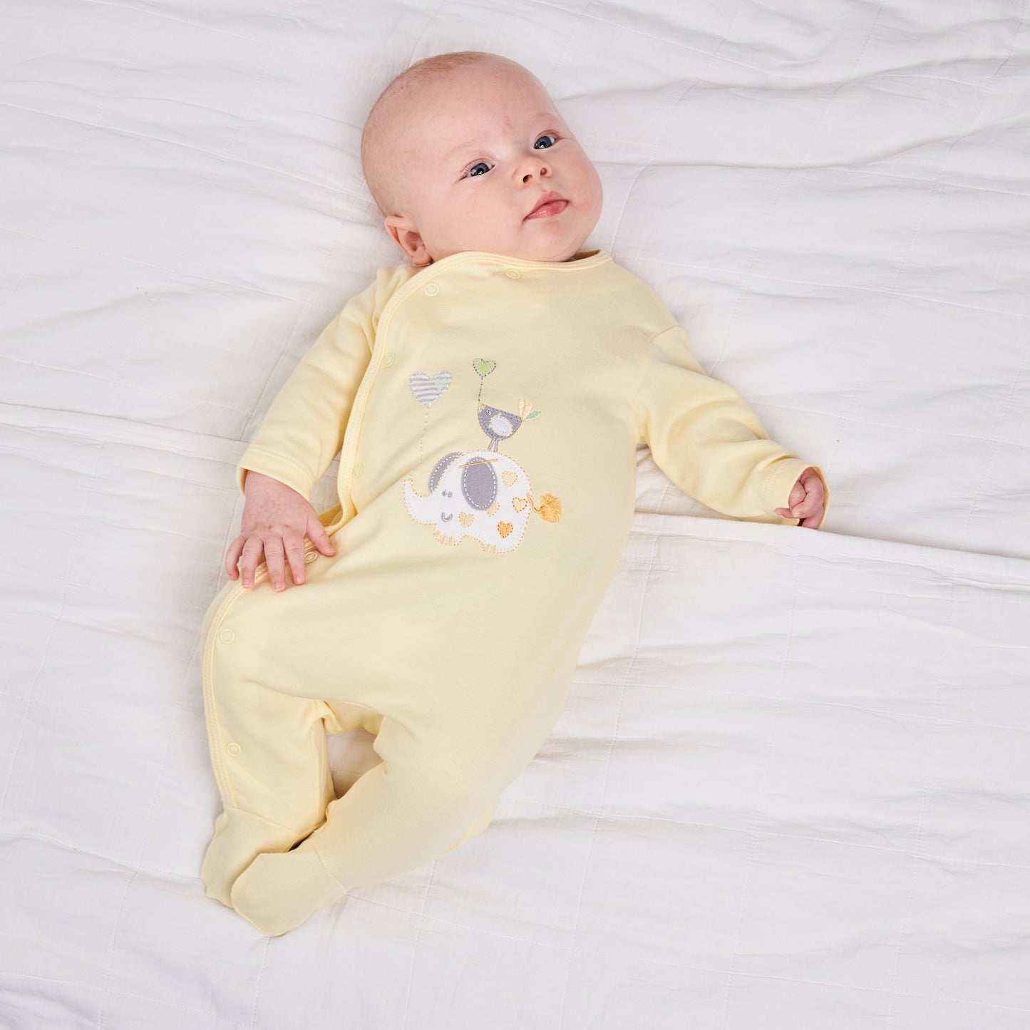 Baby in yellow onesie lying on white bedding. ELEPHANT & BIRD WITH BALLOON COTTON BABY SLEEPSUIT - LEMON YELLOW | DANDELION