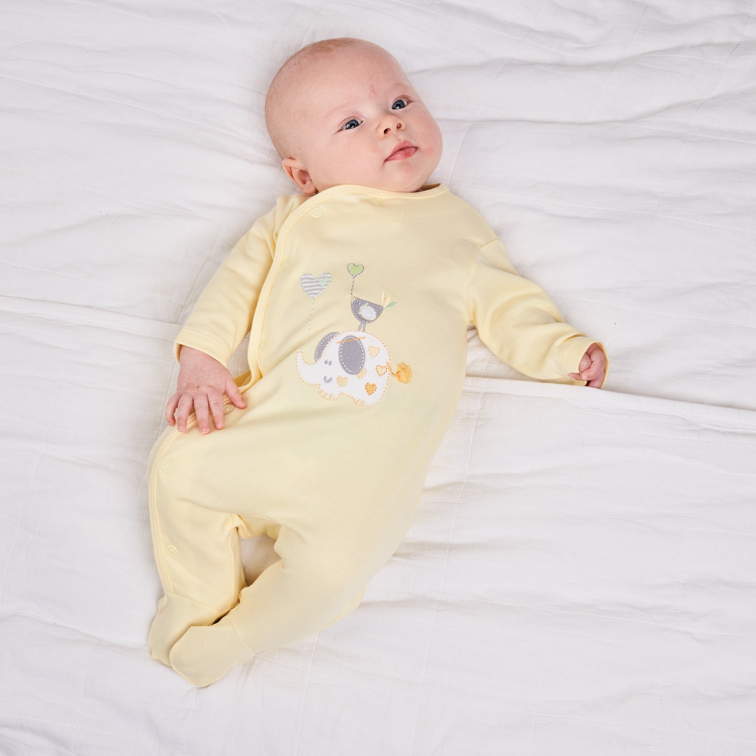 Baby in yellow onesie lying on white bedding. ELEPHANT & BIRD WITH BALLOON COTTON BABY SLEEPSUIT - LEMON YELLOW | DANDELION