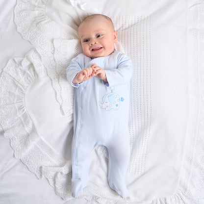 A baby lying on a white textured surface wearing a blue cotton sleepsuit with a dandelion, elephant & bird with balloon design. ELEPHANT & BIRD WITH BALLOON COTTON SLEEPSUIT - BABY BLUE | DANDELION