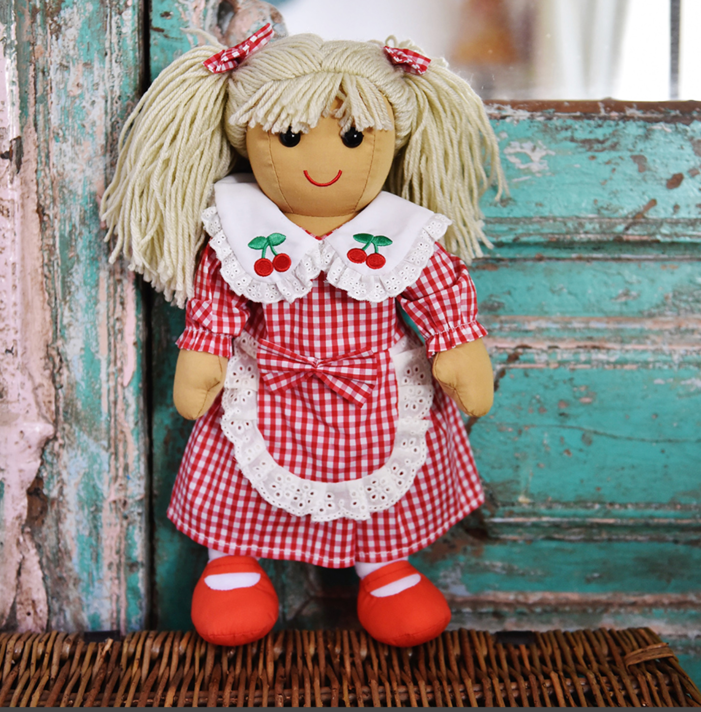 Toy doll in a red and white checkered dress with cherry details on a textured background. EMBROIDERED CHERRY RED GINGHAM RAGDOLL WITH  BLONDE HAIR AND PETER PAN COLLAR | POWELL CRAFT