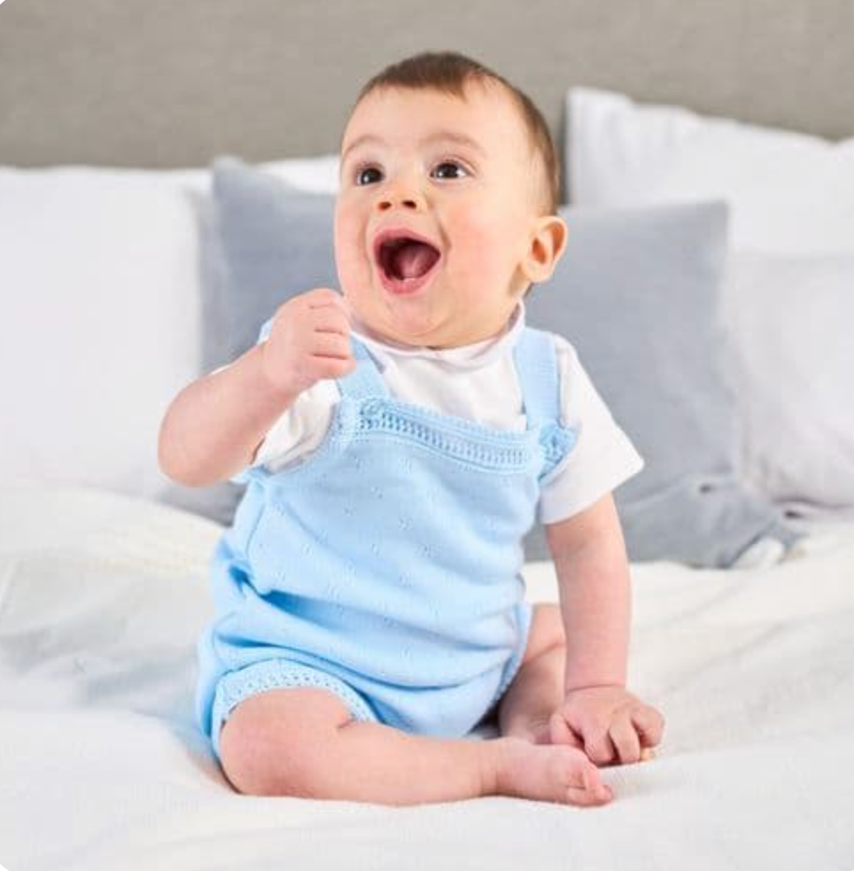 Baby in blue outfit sitting on a bed. FINE KNIT BABY ROMPER - BABY BLUE | DANDELION