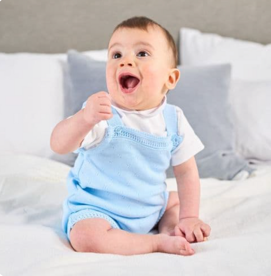 Baby in blue outfit sitting on a bed. FINE KNIT BABY ROMPER - BABY BLUE | DANDELION