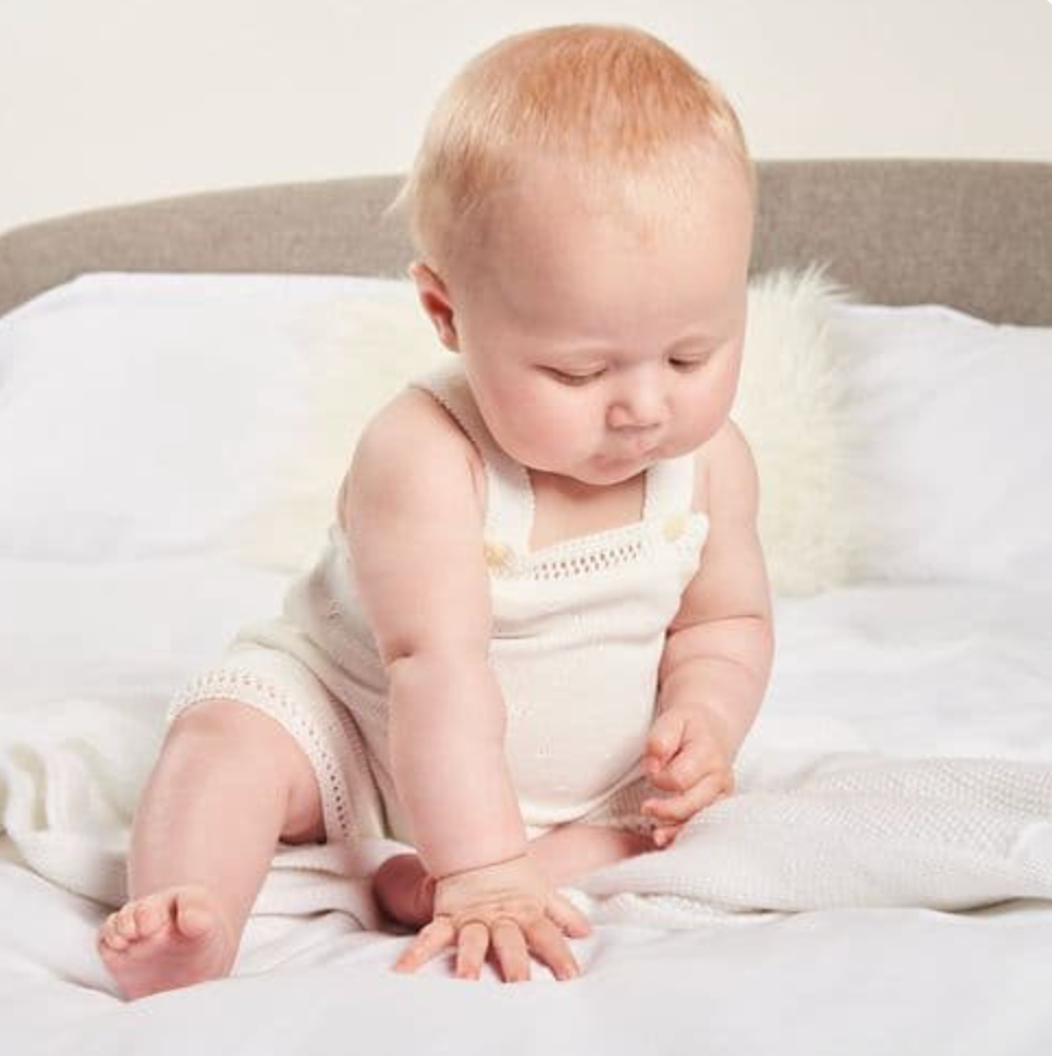 A baby sitting on a bed wearing a white knitted romper with shell button details. FINE KNIT BABY ROMPER - CLASSIC WHITE | DANDELION