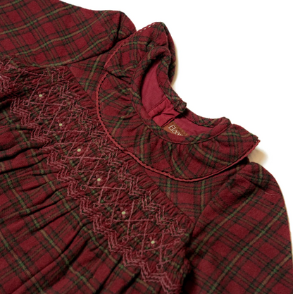 Close-up detail of the Eleanor Charles red tartan smock dress showing the hand-smocked chest yoke and ruffled collar; deep crimson and forest green tartan fabric with fine gold metallic thread in the weave clearly visible; ruffled frill collar in plain crimson fabric with picot scallop edge trim at top; full hand-smocked yoke panel below the collar showing intricate diamond lattice smocking in deep burgundy thread with tiny white French knot dots at each diamond intersection; zigzag chevron rows of smocking