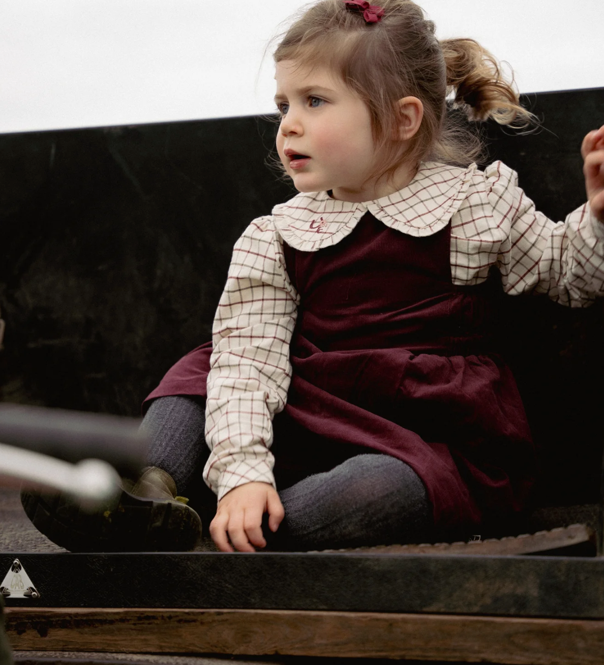 Young girl sitting inside a vintage vehicle, wearing a checkered shirt and maroon dress. GIRLS COTTON COUNTRY CHECK BLOUSE WITH PETER PAN COLLAR | ELEANOR CHARLES
