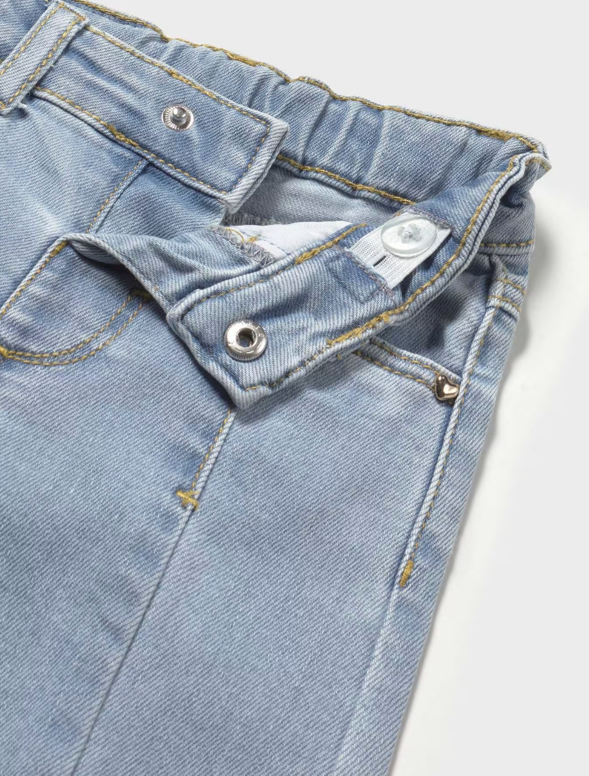 Close-up of waistband, front fastening, pocket detail and upper leg; pale ice-blue glacier wash stretch denim with weave texture and slight sheen; structured front waistband panel shown open — small round silver metal press-stud or popper snap fastening at the centre front waistband opening; white fabric internal waist adjuster tab with a small popper visible inside the waistband; elasticated gathered waistband section to the right with mustard-yellow topstitching along the edge; small gold heart-shaped met