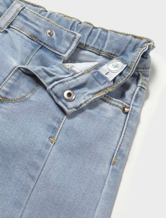 Close-up of waistband, front fastening, pocket detail and upper leg; pale ice-blue glacier wash stretch denim with weave texture and slight sheen; structured front waistband panel shown open — small round silver metal press-stud or popper snap fastening at the centre front waistband opening; white fabric internal waist adjuster tab with a small popper visible inside the waistband; elasticated gathered waistband section to the right with mustard-yellow topstitching along the edge; small gold heart-shaped met