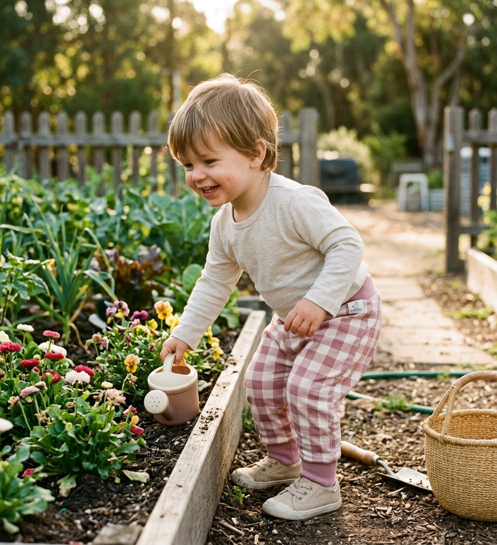 oddler, approximately 18 months–2 years, in a sunny kitchen garden; leaning over a raised wooden flower bed holding a small clay watering can; gingham trousers fully shown — large-scale dusty rose/cream check, deep ribbed waistband, ribbed ankle cuffs, and Pigeon Organics label visible on the hip; cream ribbed long-sleeve top tucked in; pansies and daisies in the garden bed; wicker basket, green hose; warm golden late afternoon light.