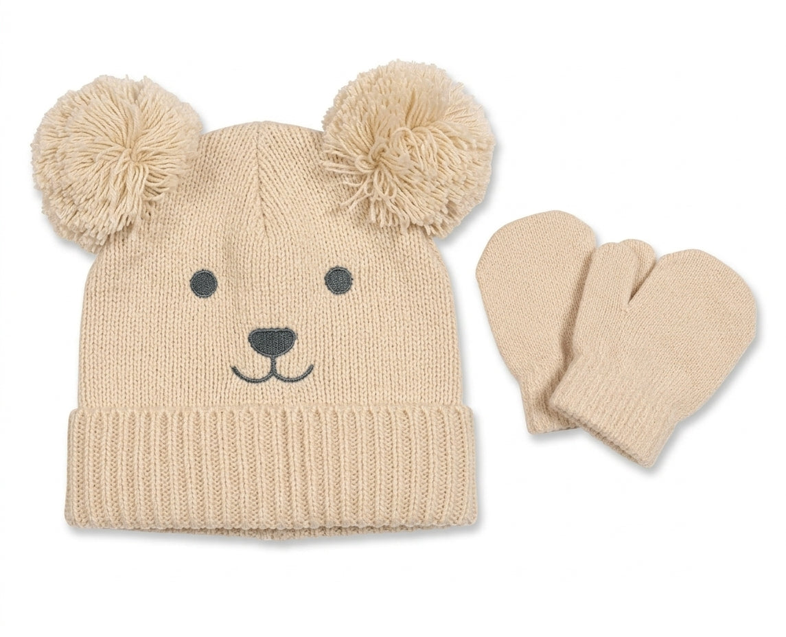 nuggle Baby Teddy Knit Hat and Mittens Set in oatmeal-cream shown flat on a white background; smooth knit beanie with a wide ribbed fold-up brim and two large fluffy pom-pom ears giving a teddy bear appearance; embroidered teddy bear face on the front with two small dark charcoal dot eyes, a charcoal triangular nose and a curved embroidered smile; matching plain oatmeal-cream knit mittens with a neat ribbed cuff and no thumb separation