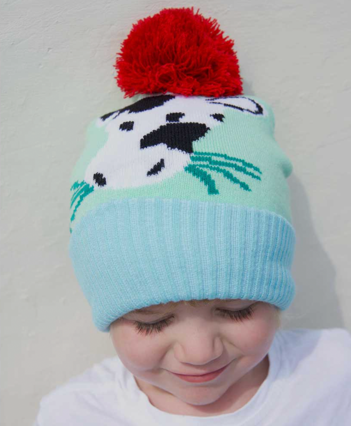 Child wearing a colorful knit hat with a COW design and red pom-pom. KID'S COW FARMYARD KNIT HAT WIHT POM-POM | POWELL CRAFT