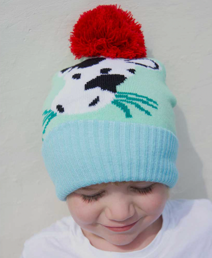Child wearing a colorful knit hat with a COW design and red pom-pom. KID'S COW FARMYARD KNIT HAT WIHT POM-POM | POWELL CRAFT