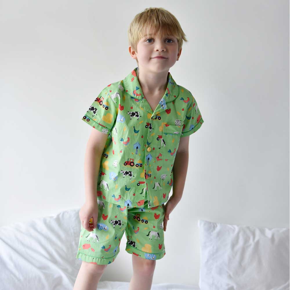 A child wearing a green pyjama set with a farmyard print, consisting of shorts and a top with short sleeves. KID'S FARMYARD SHORTS & TOP PYJAMA SET - 3 SIZES | POWELL CRAFT