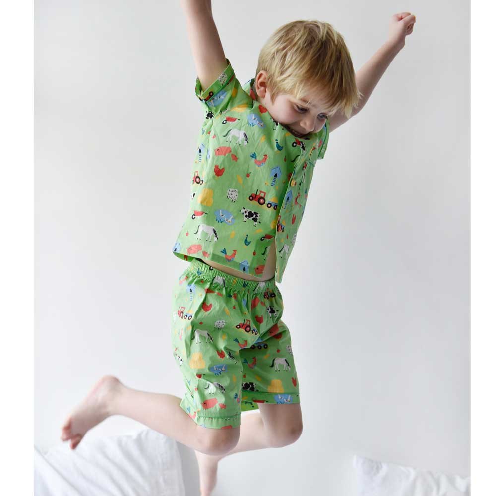 Child wearing a green outfit with vehicle prints, jumping on a bed. KID'S FARMYARD SHORTS & TOP PYJAMA SET - 3 SIZES | POWELL CRAFT