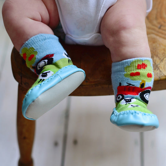 A pair of colorful knitted moccasin slipper socks with a tractor design, worn by a baby sitting on a chair. KID'S TRACTOR MOCCASIN NON-SLIP SLIPPER SOCKS - 4 SIZES | POWELL CRAFT