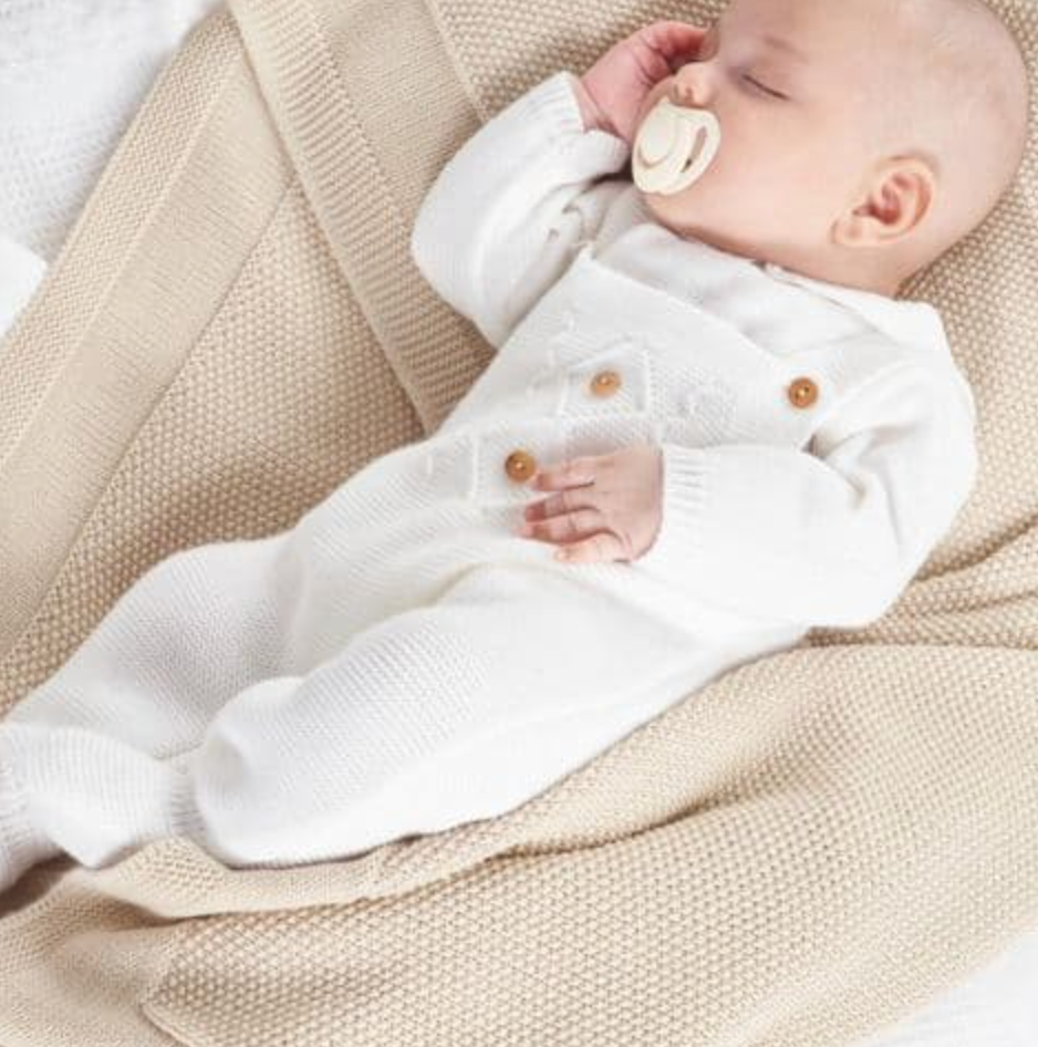 Baby in a white knitted outfit lying on a beige blanket. KNITTED BABY DUNGAREE & TOP - WHITE | DANDELION