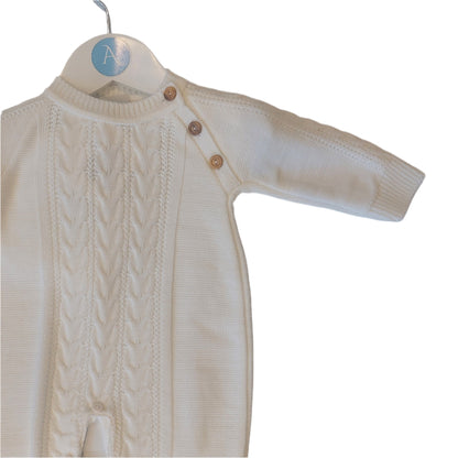 Upper body of the white knitted onesie on an Annabel's of Cranbrook branded hanger; central cable-knit chest panel, smoother knit side panels, and three round wooden shoulder buttons visible