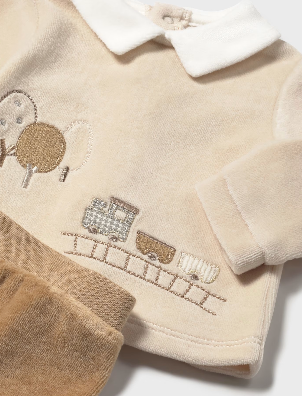 Mayoral Baby Cosy Jumper and Footed Trouser Set in Toffee — close-up detail shot on white background; angled overhead view of the front of the cream/oat velour jumper showing the embroidered and appliqué steam train scene in close detail; a little steam train engine in grey-blue and taupe tones on a stitched railway track; two carriages in taupe and cream following behind; two round-topped trees in taupe with a small circular appliqué canopy on the left; all worked in embroidery and appliqué on the soft cre
