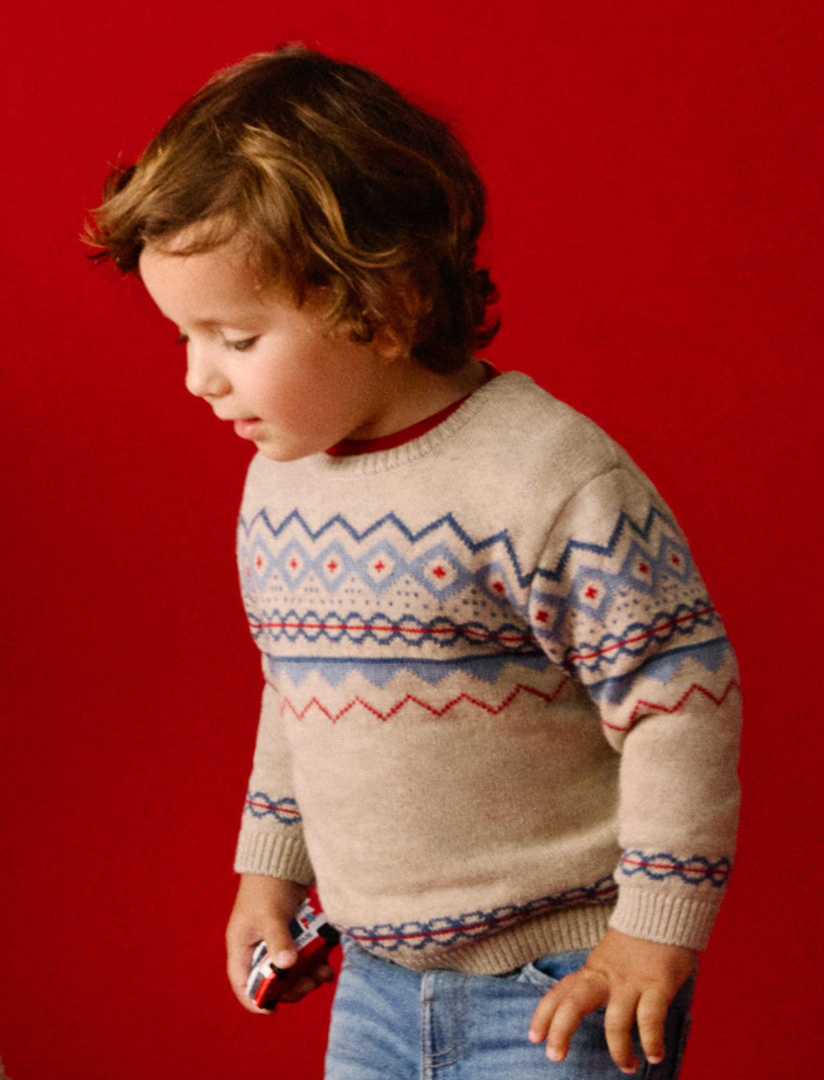 Mayoral Children's Classic Jacquard Jumper in Oat — lifestyle shot against a bold red background; a young child with curly brown hair looks downward while wearing the oat heather jumper; the classic Fair Isle-style jacquard yoke pattern in periwinkle blue, red and cream is clearly visible across the chest and sleeves; the child wears the jumper over a red crew-neck top with light blue jeans; a small toy car is held in the right hand; demonstrates the jumper worn on a child, the real-world fit and scale, and