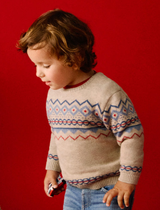 Mayoral Children's Classic Jacquard Jumper in Oat — lifestyle shot against a bold red background; a young child with curly brown hair looks downward while wearing the oat heather jumper; the classic Fair Isle-style jacquard yoke pattern in periwinkle blue, red and cream is clearly visible across the chest and sleeves; the child wears the jumper over a red crew-neck top with light blue jeans; a small toy car is held in the right hand; demonstrates the jumper worn on a child, the real-world fit and scale, and