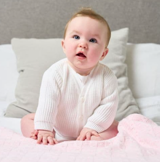 Baby wearing a white cardigan sitting on a pink blanket with a neutral background. PREMATURE BABY CARDIGAN RIBBED KNIT - CLASSIC WHITE | DANDELION
