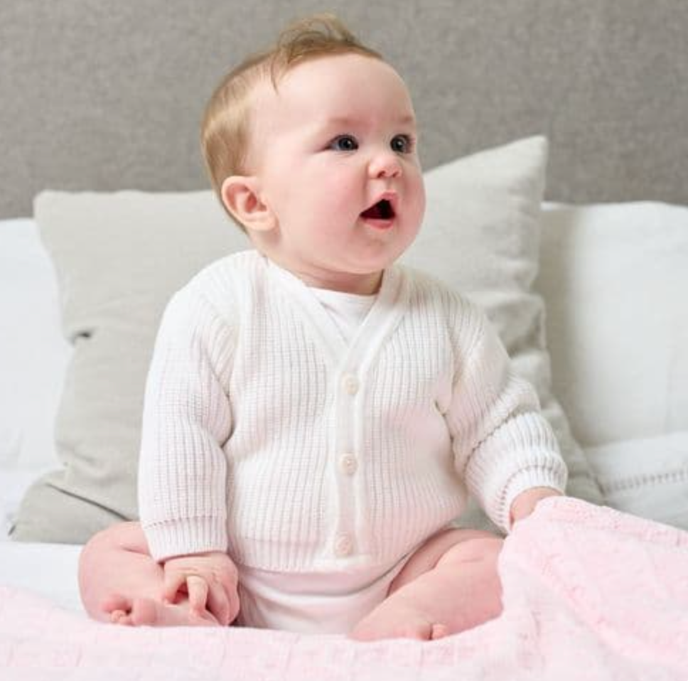 Baby wearing a white ribbed cardigan sitting on a bed with light gray pillows. PREMATURE BABY CARDIGAN RIBBED KNIT - CLASSIC WHITE | DANDELION
