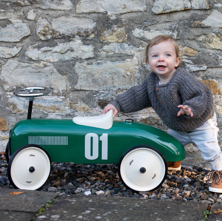 Child playing with a green toy car against a stone wall. RIDE ON CLASSIC CAR - BRITISH RACING GREEN WITH WHITE SEAT | VILAC 