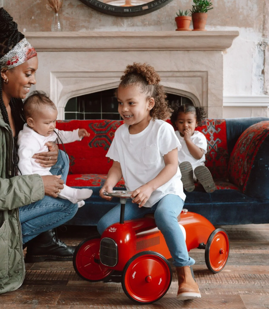 Woman with two children on a red couch, one child riding a red toy car. RIDE ON CLASSIC CAR - RED WITH RED WHEELS & TAN SEAT | VILAC