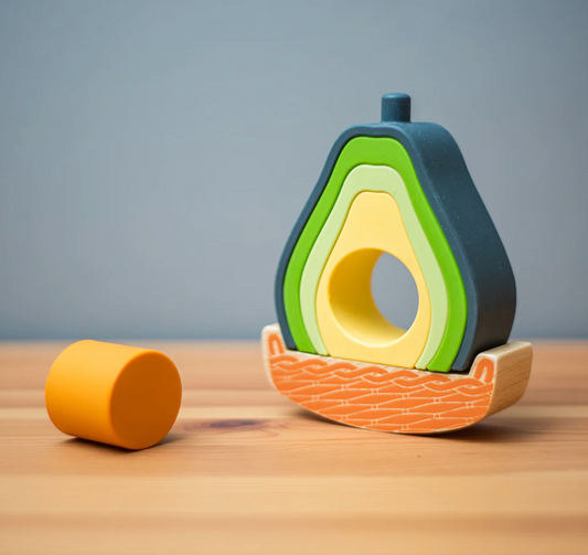 Bigjigs avocado-shaped silicone stacking toy displayed on a wooden surface beside its curved FSC® wood rocking tray, showing how the green nesting pieces can be separated and rearranged for open-ended sensory play.