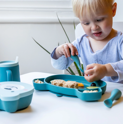 Child using a blue sippy cup and eating from a divided plate with utensils on a white surface. SILICONE BABY TRAVEL CUTLERY SET WITH CASE - 2 COLOURS | TUM TUM