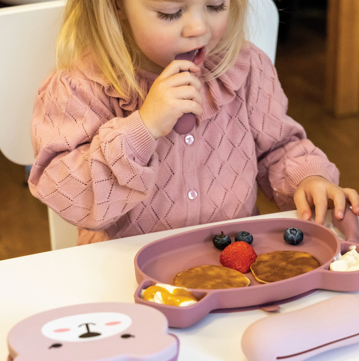 Child eating from a pink plate with food at a dining table. SILICONE TRAVEL WEANING BOWL & SPOON SET - 2 COLOURS | TUM TUM 