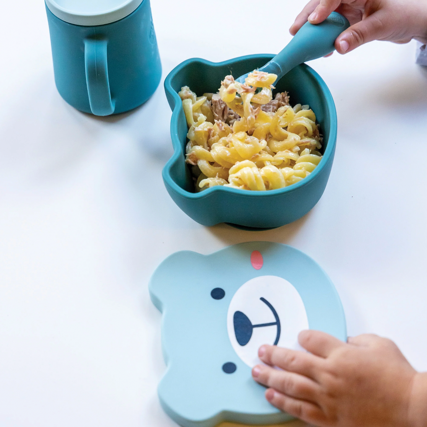 Children's blue bowl with pasta and a spoon, next to a matching placemat with a bear face design. SILICONE TRAVEL WEANING BOWL & SPOON SET - 2 COLOURS | TUM TUM 