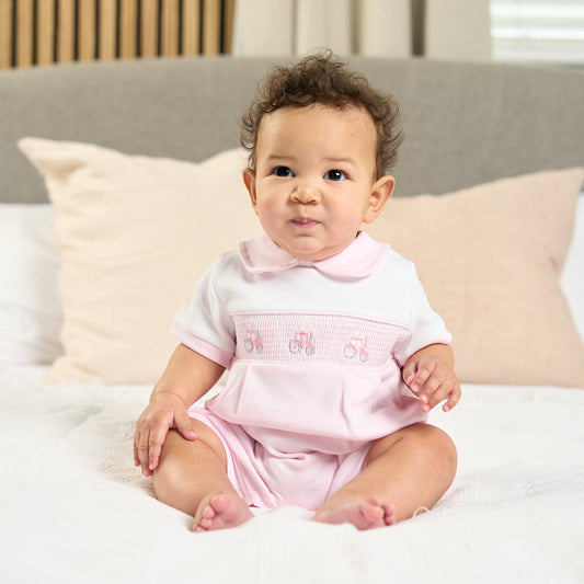 A baby sitting on a bed wearing a pink romper with a tractor and name smocked on the front, and a Peter Pan collar. SMOCKED TRACTOR COTTON SUMMER ROMPER OUTFIT - BABY PINK | DANDELION
